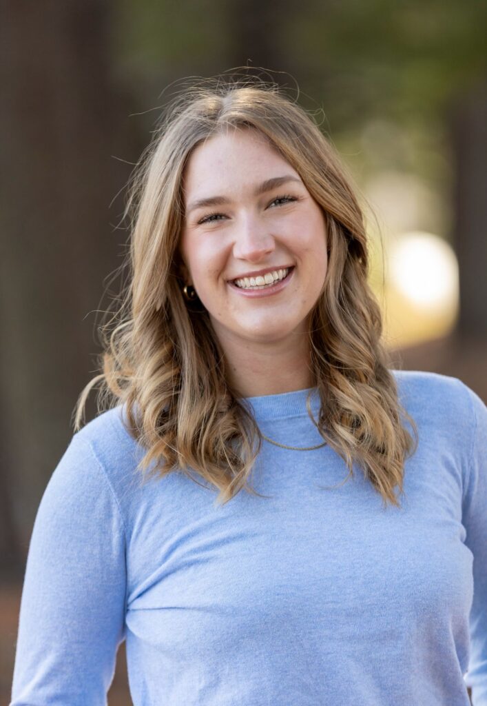 A woman with wavy blonde hair smiles outdoors, wearing a light blue long-sleeve shirt.