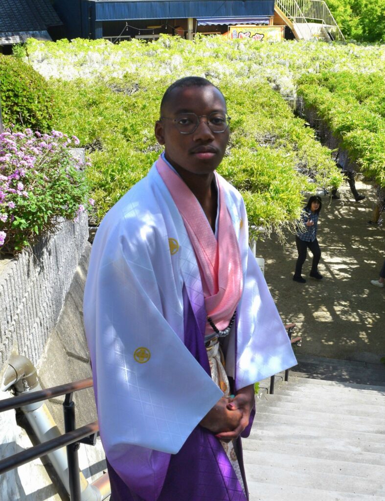 A person in a white and purple kimono stands on outdoor steps with greenery and other people in the background.