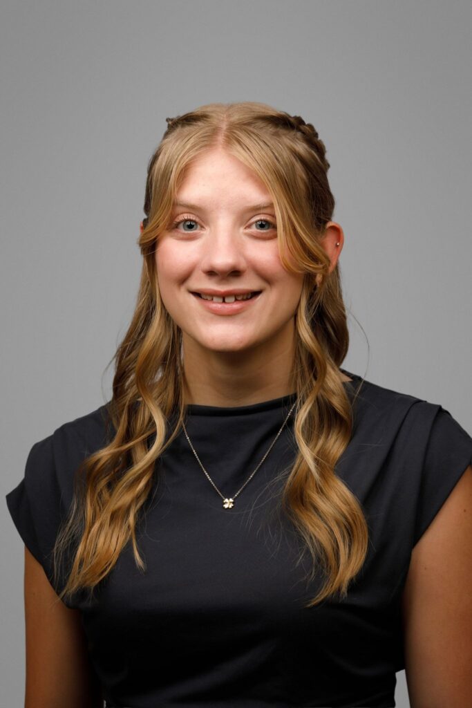 Young woman with long, wavy blonde hair wearing a dark top and a necklace, smiling at the camera against a gray background.