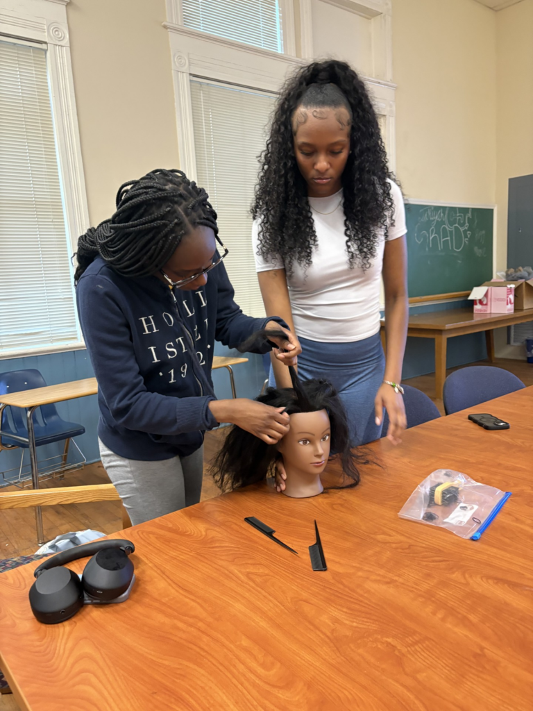 Two young women style a mannequin head with hair tools at a table in a classroom setting.