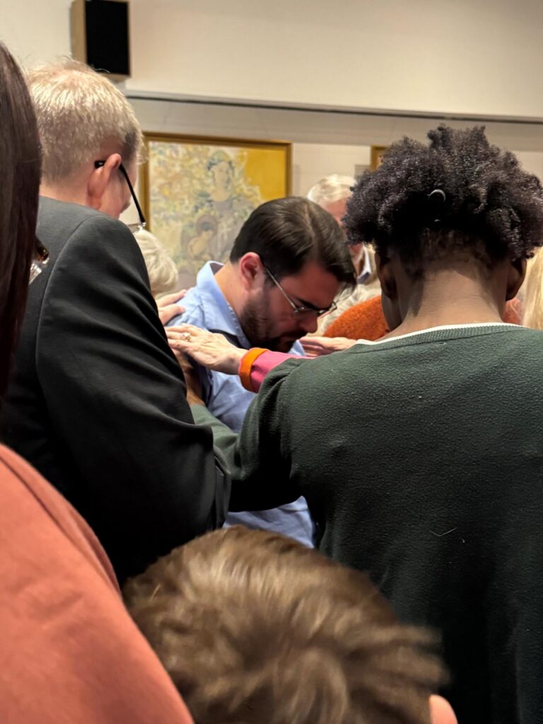 Several people stand in a group, placing their hands on a man with glasses who has his head bowed.