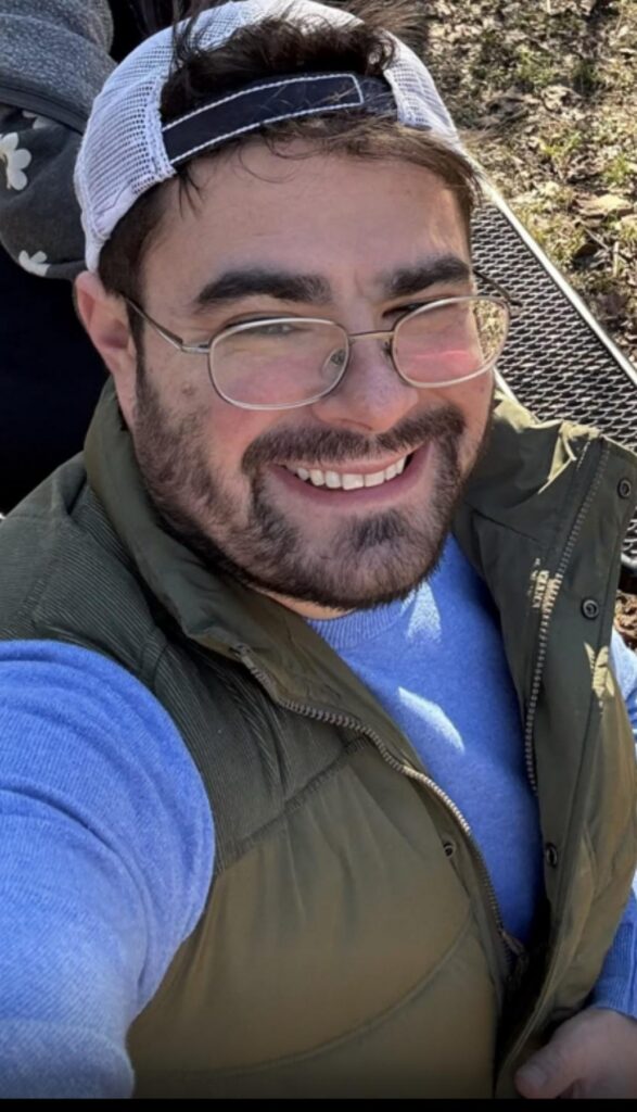 Man with glasses and a cap smiles outdoors, wearing a blue shirt and green vest. Sunlight and grass in the background.
