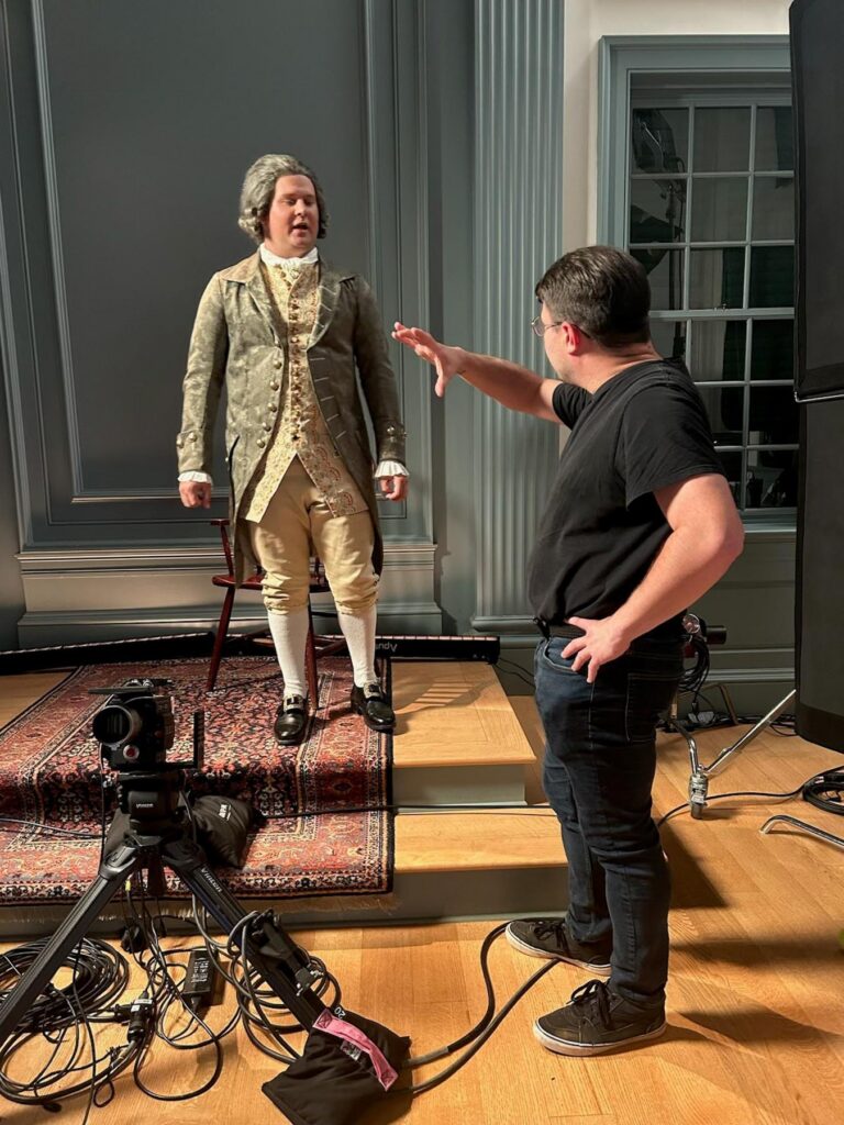 A person in 18th-century costume stands on a platform as another person gives directions beside filming equipment.