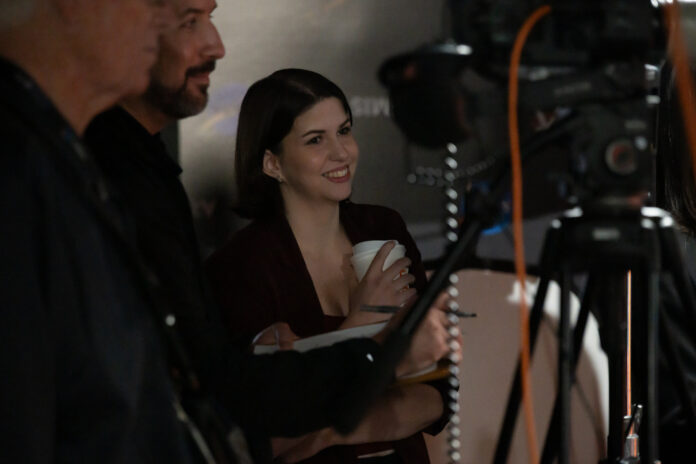 jsc2025e080727-2 Katie Konans smiles while standing behind a camera setup with two other people at an indoor event.