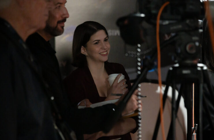 Katie Konans smiles while standing behind a camera setup with two other people at an indoor event.