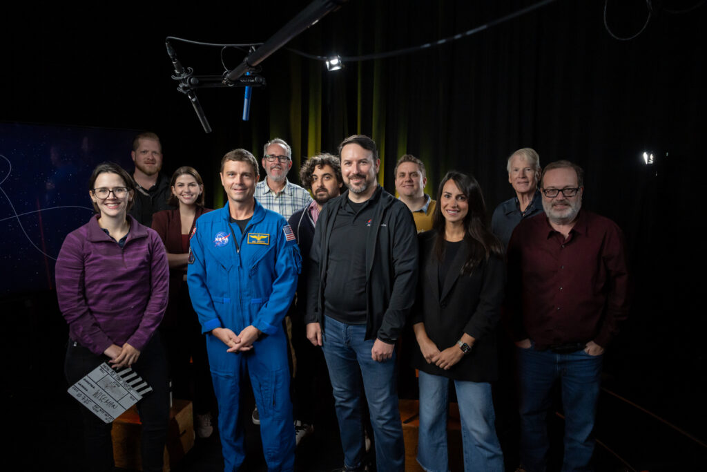 A group of eleven people, including one in a NASA flight suit, pose together in a studio with filming equipment visible.