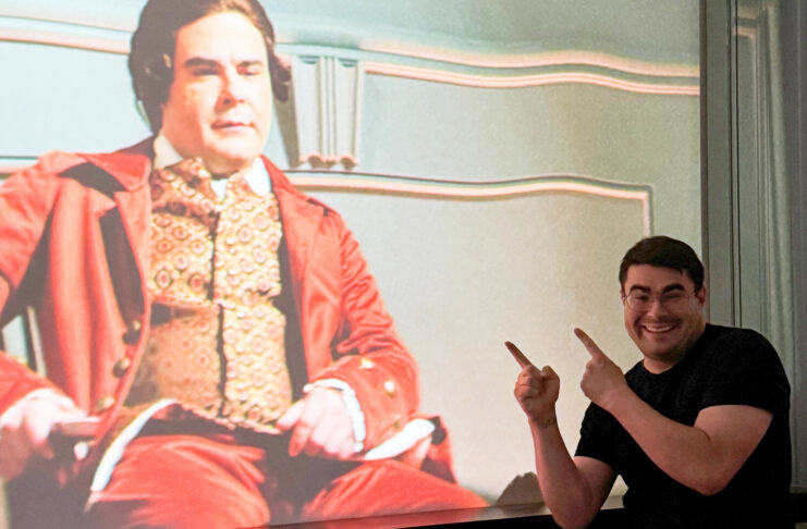 Smiling man points at a projected image of an actor in historical costume on a screen.