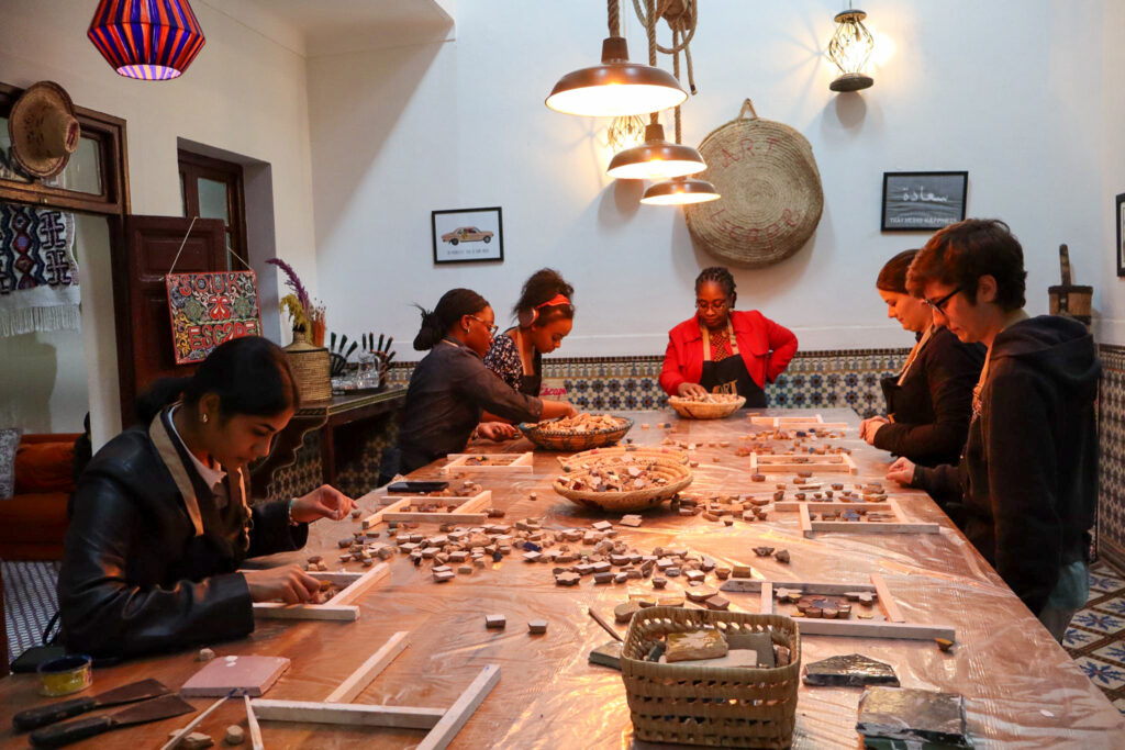Six people sit around a table assembling mosaic pieces in a well-lit, decorated room with patterned walls.