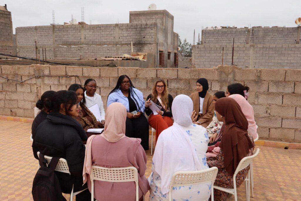 A group of women sit in a circle on chairs, having a discussion outside near unfinished concrete buildings.