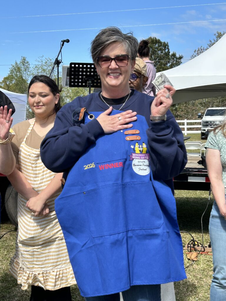Smiling woman wearing a blue apron labeled 2024 WINNER holds cash while standing outdoors among others at an event.
