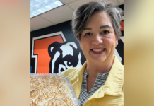 Staff member wins banana pudding contest at state festival A person in a yellow jacket holds a cake and smiles, standing in front of a wall with a bulldog graphic.