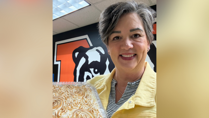 A person in a yellow jacket holds a cake and smiles, standing in front of a wall with a bulldog graphic.