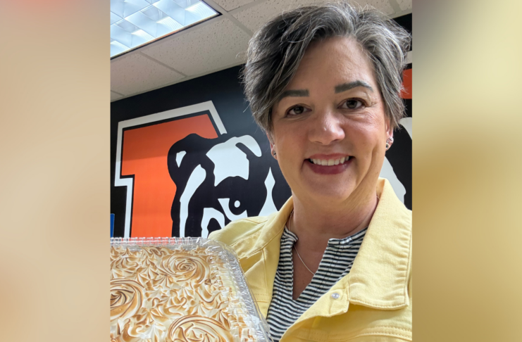 A person in a yellow jacket holds a cake and smiles, standing in front of a wall with a bulldog graphic.