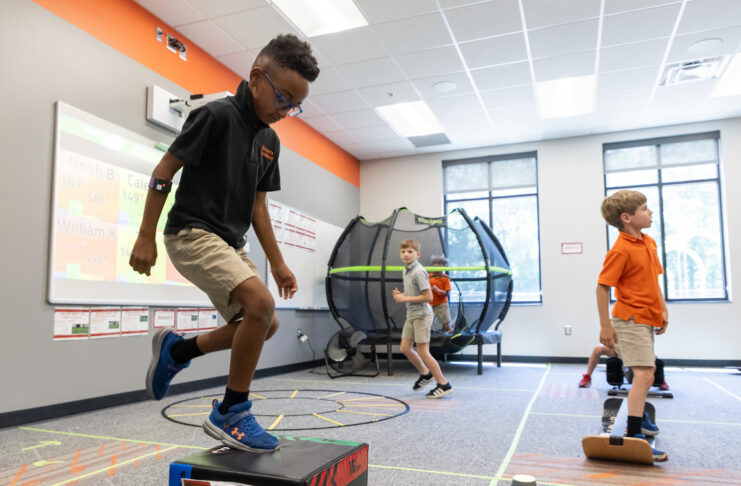 Three boys participate in physical activities in a classroom with exercise equipment and a trampoline in the background.