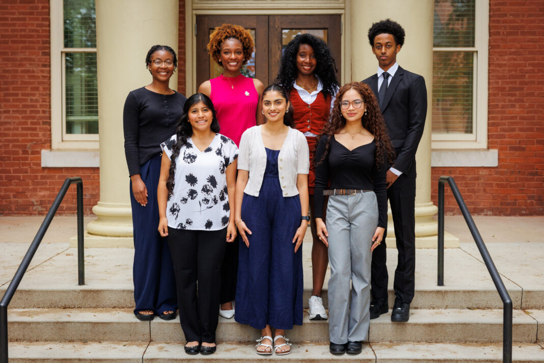 Seven people stand in two rows on steps outside a brick building with columns, facing the camera and smiling.