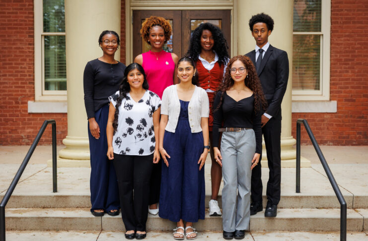Seven people stand in two rows on steps outside a brick building with columns, facing the camera and smiling.
