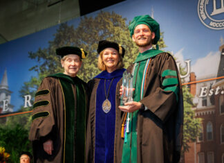 Outstanding School of Medicine graduates and faculty honored during 2026 commencement Three people in academic regalia pose on stage at a graduation ceremony; one holds a glass award and smiles.