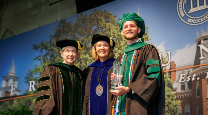 Outstanding School of Medicine graduates and faculty honored during 2026 commencement Three people in academic regalia pose on stage at a graduation ceremony; one holds a glass award and smiles.