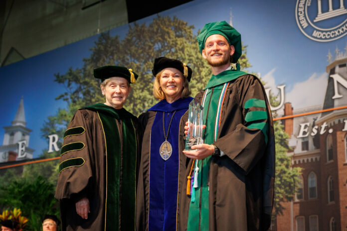 Three people in academic regalia pose on stage at a graduation ceremony; one holds a glass award and smiles.
