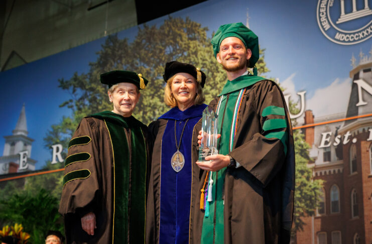 Three people in academic regalia pose on stage at a graduation ceremony; one holds a glass award and smiles.