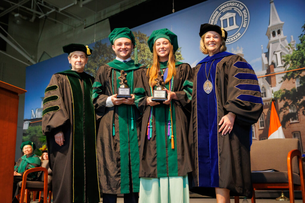 Four people in academic regalia pose at a graduation ceremony; two hold trophies and smile at the camera.
