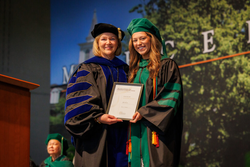 Two women in academic regalia pose together on stage holding a certificate at a graduation ceremony.