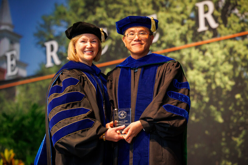 Two people in academic regalia smile and hold an award at a graduation or academic ceremony.