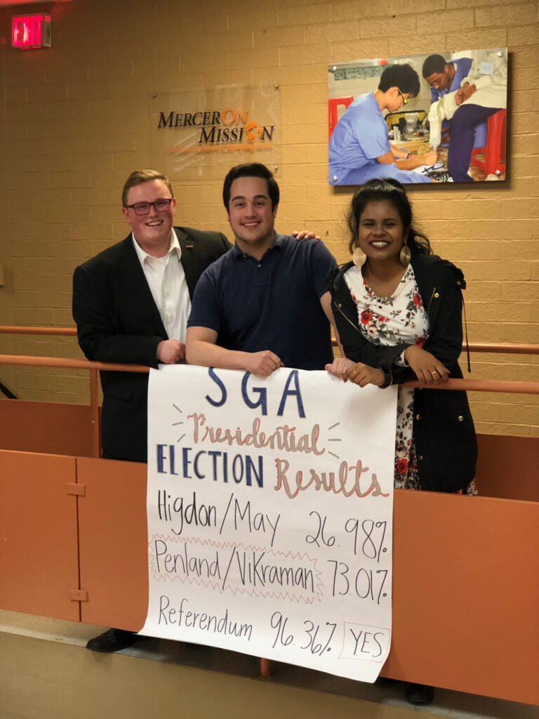 Three people stand indoors holding a sign showing student government election results at Mercer University.