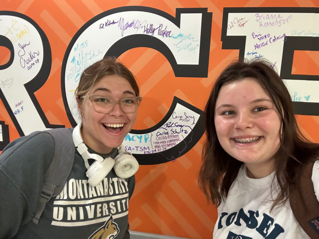 Two smiling students pose in front of an orange wall covered in signatures and messages.