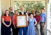 Family who grew up in Mercer CAPS house shares its 136-year history Nine people stand on a porch, with one man in the center holding a framed document and photo.