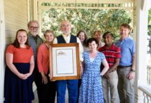 Family who grew up in Mercer CAPS house shares its 136-year history Nine people stand on a porch, with one man in the center holding a framed document and photo.