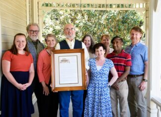 Family who grew up in Mercer CAPS house shares its 136-year history Nine people stand on a porch, with one man in the center holding a framed document and photo.