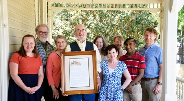 Family who grew up in Mercer CAPS house shares its 136-year history Nine people stand on a porch, with one man in the center holding a framed document and photo.