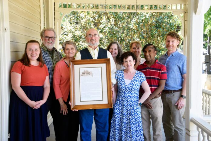 Nine people stand on a porch, with one man in the center holding a framed document and photo.