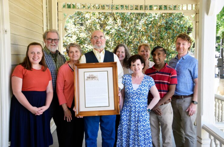 Nine people stand on a porch, with one man in the center holding a framed document and photo.