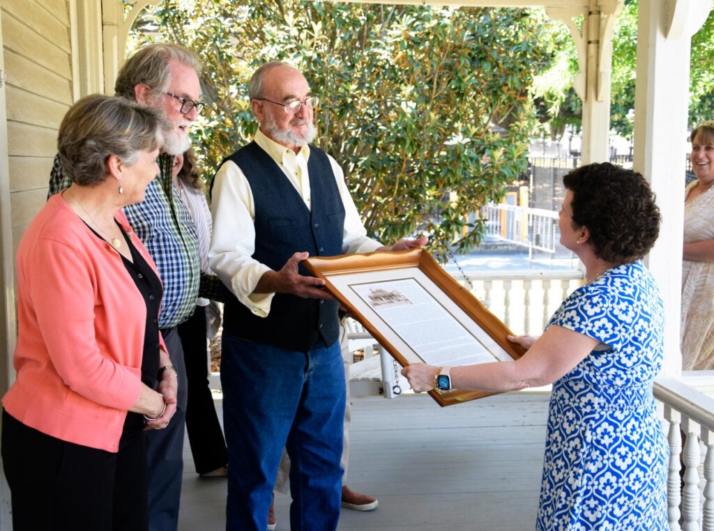 A woman in a blue dress presents a framed document to three people standing on a porch during a daytime gathering.
