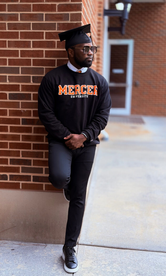 A man in a graduation cap and Mercer University sweatshirt leans against a brick wall outdoors.