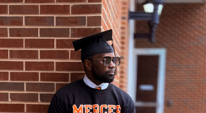 Graduate Spotlight: Deon Brown A man in a graduation cap and Mercer University sweatshirt leans against a brick wall outdoors.
