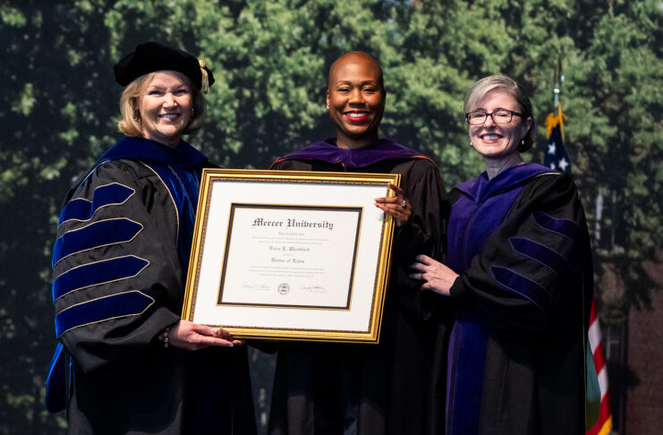 Three women in academic regalia hold a framed Mercer University diploma at a graduation ceremony.
