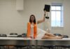 Graduate Spotlight: Virginia Fox A woman in graduation attire poses on a table, holding a cap, with a skeleton model in the background.
