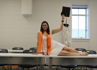 Graduate Spotlight: Virginia Fox A woman in graduation attire poses on a table, holding a cap, with a skeleton model in the background.