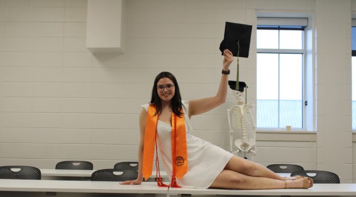 Graduate Spotlight: Virginia Fox A woman in graduation attire poses on a table, holding a cap, with a skeleton model in the background.