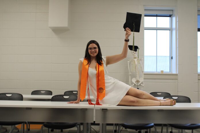 A woman in graduation attire poses on a table, holding a cap, with a skeleton model in the background.