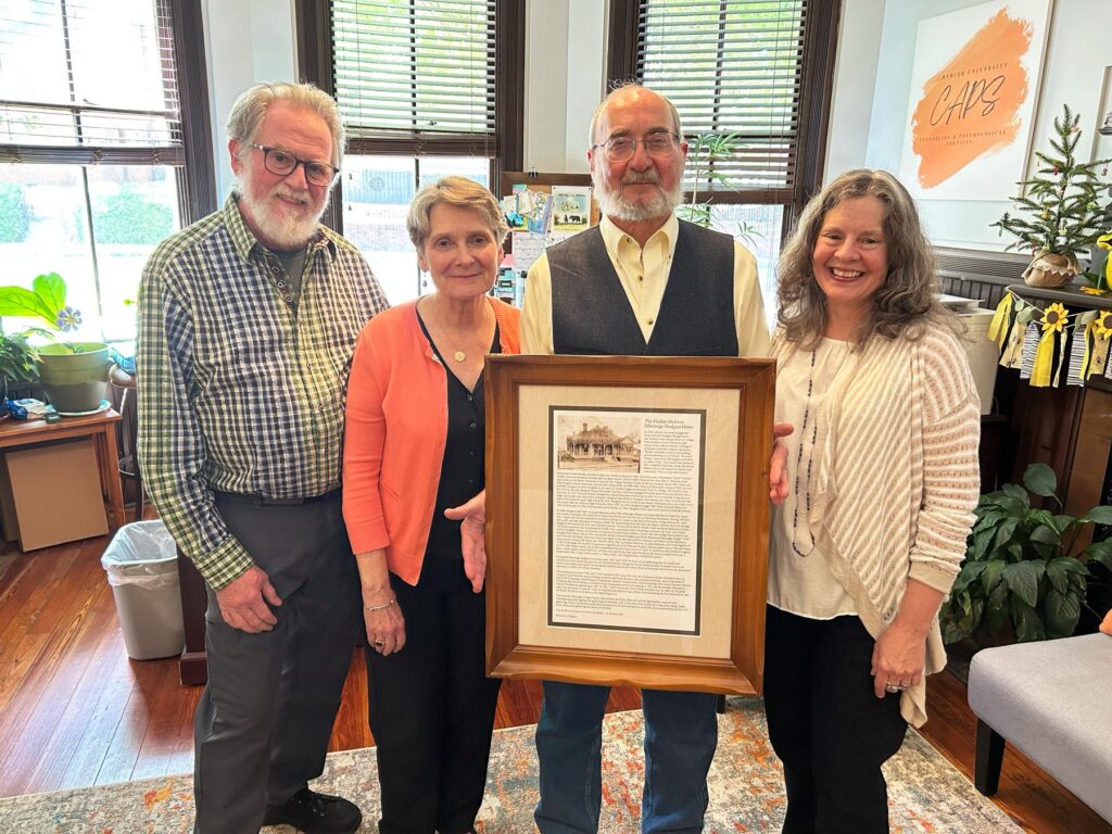 Four adults stand indoors, smiling, while one person holds a framed document with a photo and text.