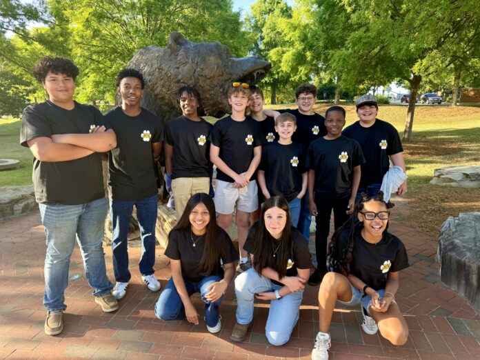 A group of students in matching black shirts poses in front of a bear statue outdoors on a sunny day.