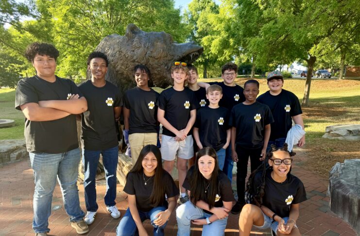 A group of students in matching black shirts poses in front of a bear statue outdoors on a sunny day.