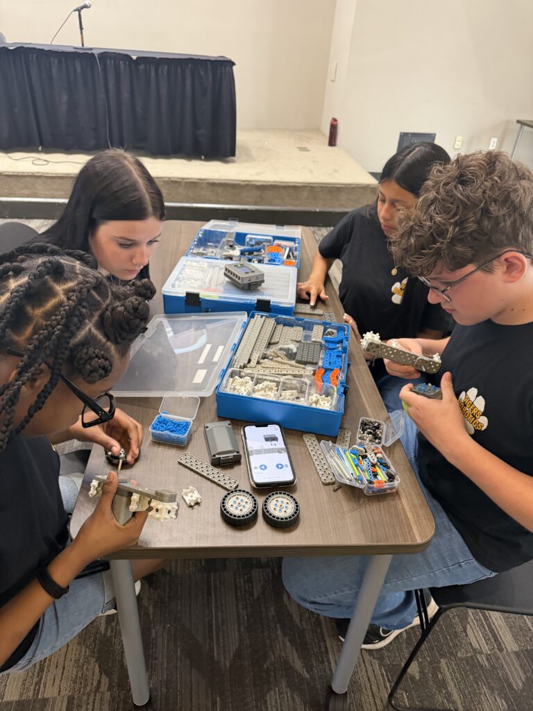 Four students sit around a table assembling robotics kits with various parts and tools spread out in front of them.