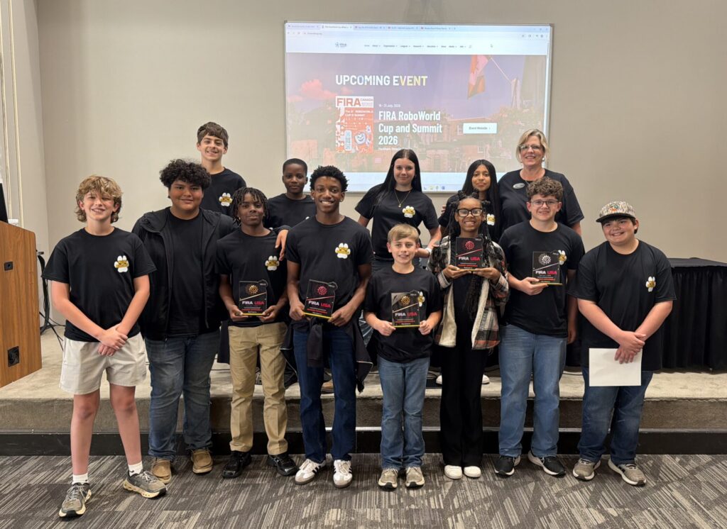 A group of students and an adult pose indoors, holding awards and wearing matching black shirts.