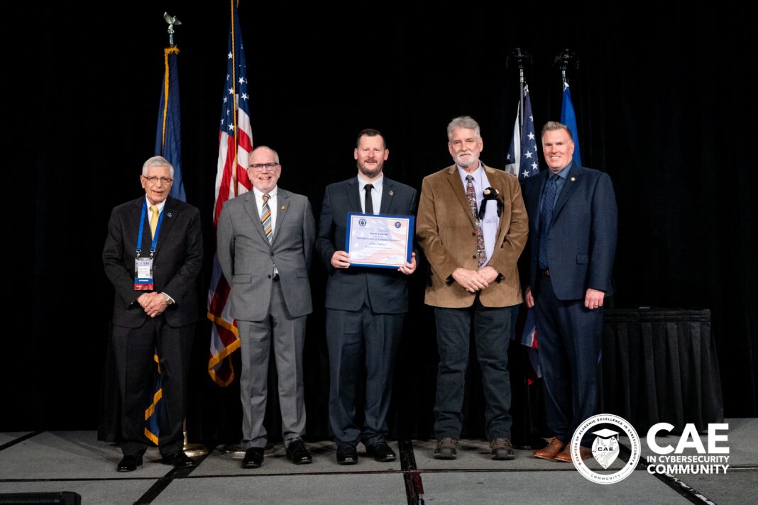 Five men in suits stand on stage with flags, one holding a certificate; CAE in Cybersecurity Community logo is visible.