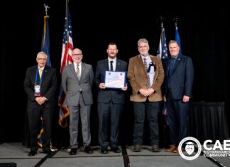 Mercer formally receives NSA Center of Academic Excellence designation Five men in suits stand on stage with flags, one holding a certificate; CAE in Cybersecurity Community logo is visible.